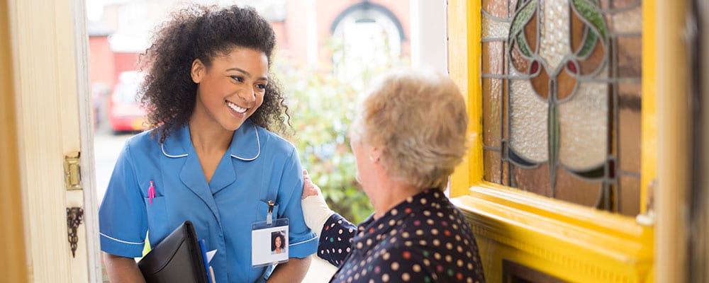A nurse visits her patient at home and smiles as she's welcomed through the door.