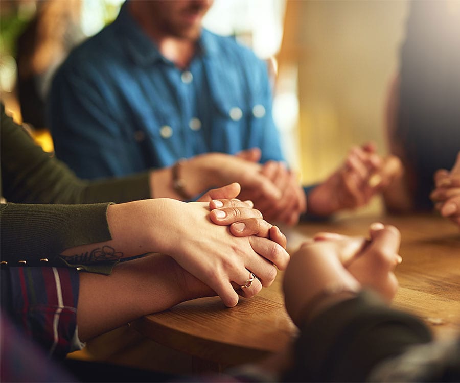 A group of people around a table hold hands and lower their heads in prayer.