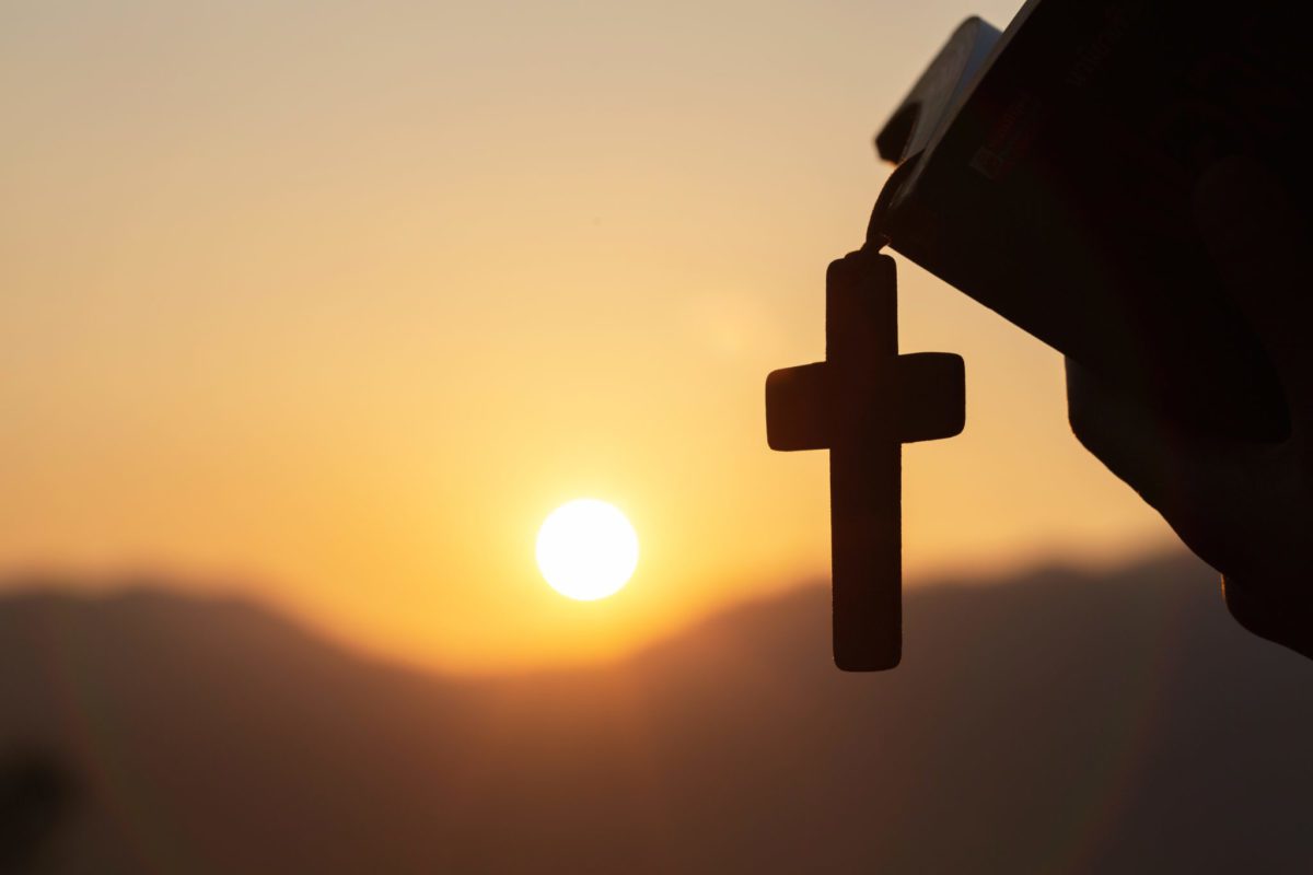 A cross hangs from a Bible in front of a sunset.